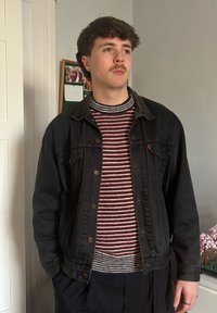 Tânăr cu mustață, purtând un pulover cu dungi și o jachetă din denim negru, stând în interior lângă un perete și o fotografie înrămată.