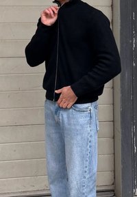 Άτομο που φοράει μαύρο πουλόβερ με φερμουάρ και ανοιχτόχρωμα τζιν, στέκεται μπροστά σε μια μπεζ τοιχοποιία με πάνελ.