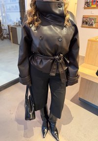 Personne portant une veste en cuir noire ceinturée, un pantalon noir, des bottes noires pointues et tenant un sac à main noir à l'intérieur.