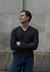 Schwarz-weiß gestreiftes Langarmshirt mit V-Ausschnitt, kombiniert mit blauen Jeans. Im Hintergrund ist eine strukturierte Steinmauer zu sehen.