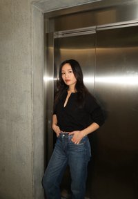 Black button-up sweater paired with light blue high-waisted jeans. The subject strikes a pose in front of a metallic elevator door, set against a concrete wall backdrop.