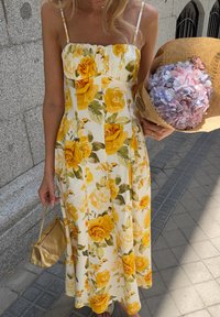 Robe midi blanche avec imprimé de roses jaunes, bretelles fines, corsage froncé et jupe évasée. Un sac à main doré et un bouquet d'hydrangeas sont tenus.
