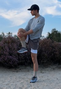 Un uomo atletico in una maglietta a maniche lunghe grigia chiara, pantaloni corti grigi e scarpe da corsa grigie si allunga su un terreno sabbioso vicino a cespugli viola.