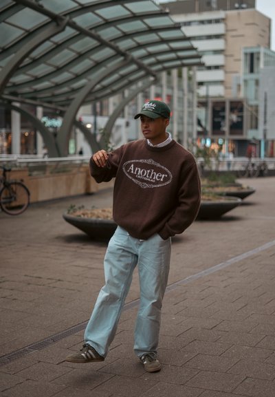 Suéter oversize marrón con el logo "Another", jeans de mezclilla azul claro y gorra verde. Zapatillas con franjas blancas. Entorno urbano con jardineras.