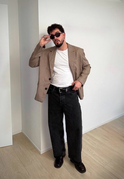 Blazer marrón de cuadros de doble botonadura sobre una camiseta blanca, combinado con jeans negros holgados y zapatos oscuros pulidos. El modelo lleva gafas de sol negras.