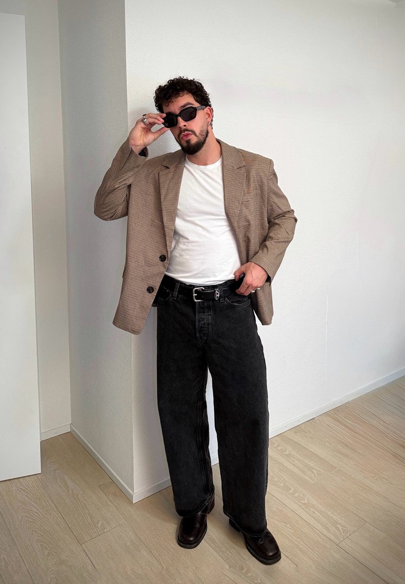 Veste croisée en tartan brun portée sur un T-shirt blanc, associée à un jean noir ample et des chaussures foncées et vernies. Le mannequin porte des lunettes de soleil noires.