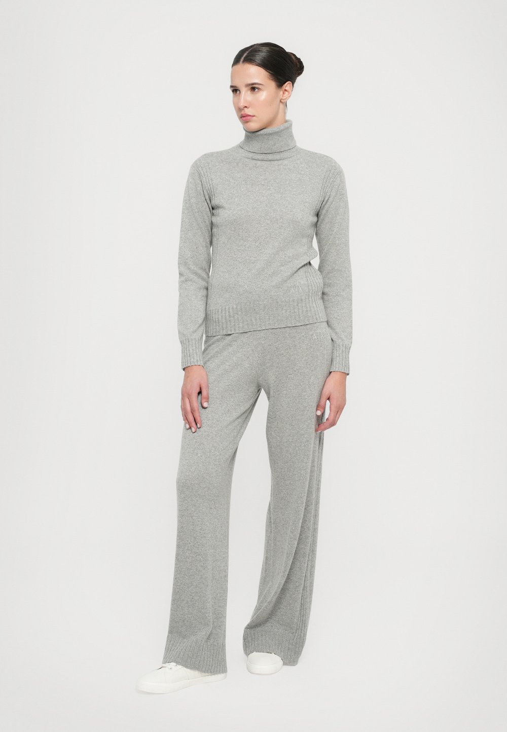 Siv pulover s turtleneckom in široke hlače, oba z rebrastim detajlom. Model nosi bele superge in stoji pred enostavno ozadje.