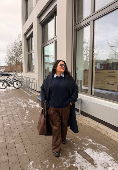 Mujer con gafas de sol caminando por una acera nevada junto a un edificio moderno con grandes ventanas, llevando un bolso tote marrón, vistiendo un abrigo azul marino y pantalones a rayas.