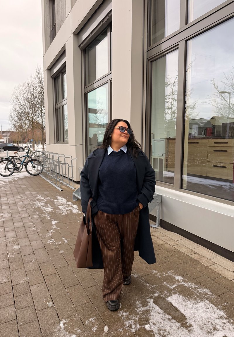 Femme en lunettes de soleil marchant sur un trottoir enneigé à côté d'un bâtiment moderne aux grandes fenêtres, portant un sac fourre-tout marron, un manteau bleu marine et un pantalon rayé.