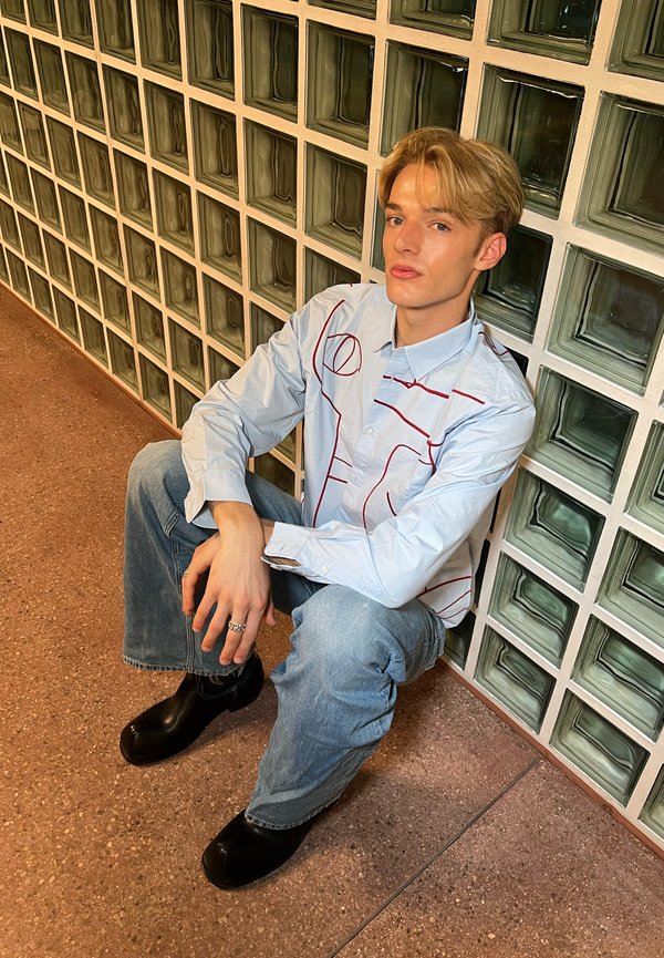 Šviesiai mėlyna marškinėliai su raudonu linijiniu raštu, derinami su šviesiais džinsais ir juodais mediniais sandalais. Fone matyti tekstūruotos stiklo plytos.