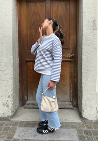 Ριγέ πουκάμισο μακρυμάνικο σε ανοιχτό μπλε και λευκό, συνδυασμένο με ανοιχτό μπλε τζιν και μαύρα αθλητικά παπούτσια. Κρατάει μία μπεζ τσάντα με σχέδιο.