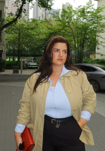 Mujer con cabello largo y oscuro que lleva una chaqueta beige sobre una camiseta azul clara y pantalones negros, sosteniendo un bolso rojo, de pie al aire libre con árboles y edificios detrás.