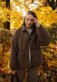 Mujer con abrigo de teddy marrón y pantalones de pana se encuentra entre hojas de otoño amarillas, tocándose suavemente el cabello con los ojos cerrados.