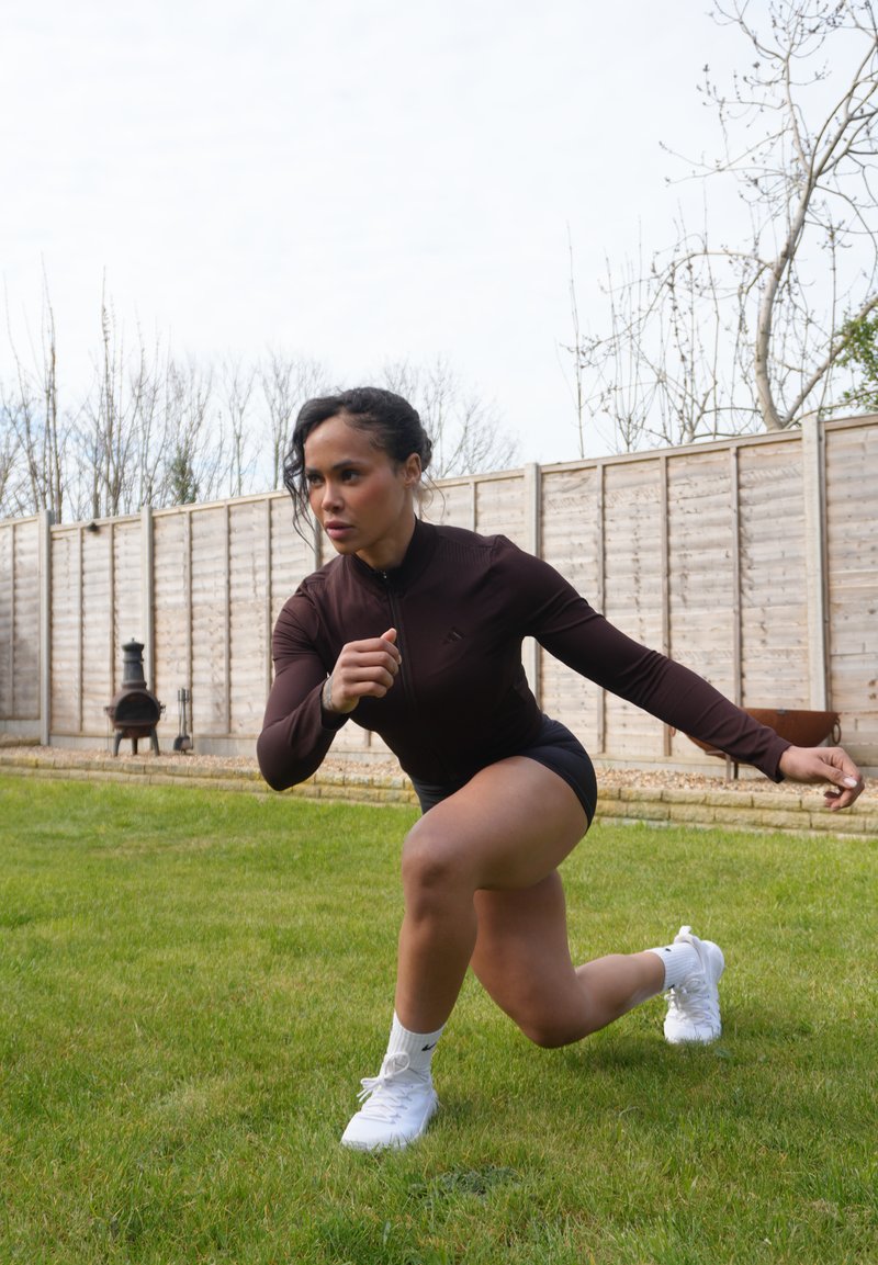 Femme en tenue de sport fait des fentes sur l'herbe dans le jardin, avec une clôture en bois et des arbres dénudés en arrière-plan.