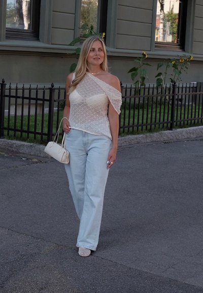 Top blanco de un solo hombro con patrón texturizado, jeans de pierna ancha azul claro y un pequeño bolso crema; combinado con zapatos blancos texturizados.