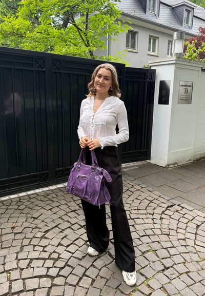 Bolso de ante morado con doble asa, detalles de hardware plateado y un bolsillo con cremallera en la parte delantera. Combinado con una blusa blanca de manga larga y pantalones negros.