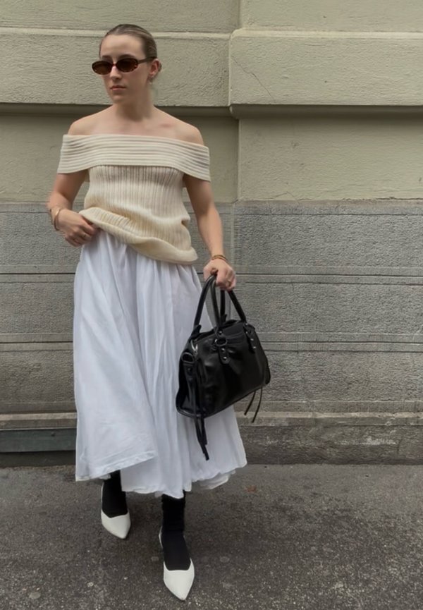 Naine kannab beežist õlgadeta kampsunit, pikka valget seelikut, musti sukkpükse, valgeid teravate ninaga kingi, tumedaid päikeseprille, hoiab käes musta käekotti.