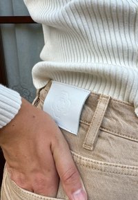 Bežiniai džinsai su baltu reljefiniu odiniu etikete nugaros pusėje. Megztinis yra baltyje, su tekstūruoto mezgimo raštu. Rankos poilsis kišenėje.