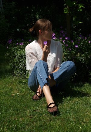 Blusa blanca bordada, jeans azules desgastados, zapatos de plataforma de malla negra. Sosteniendo una flor morada, sentada en la hierba con flores de fondo.