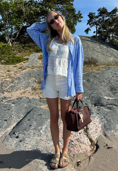 Camisa a rayas azul claro, blusa de encaje blanca, pantalones cortos blancos y bolso de cuero marrón. Sandalias oliva con correas de hebilla. Fondo de rocas y vegetación.