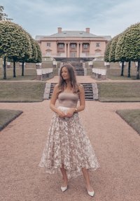 Beige tanktop, bloemenkant midi rok met een doorschijnende overlay, en witte hoge hakken; staand op een grindpad met groen en een achtergrond van een herenhuis.