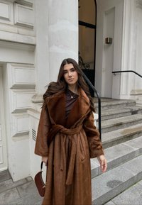 Brown faux-fur coat with wide collar and belt, layered over a dark top. Woman holding a small brown bag, standing by stone steps.
