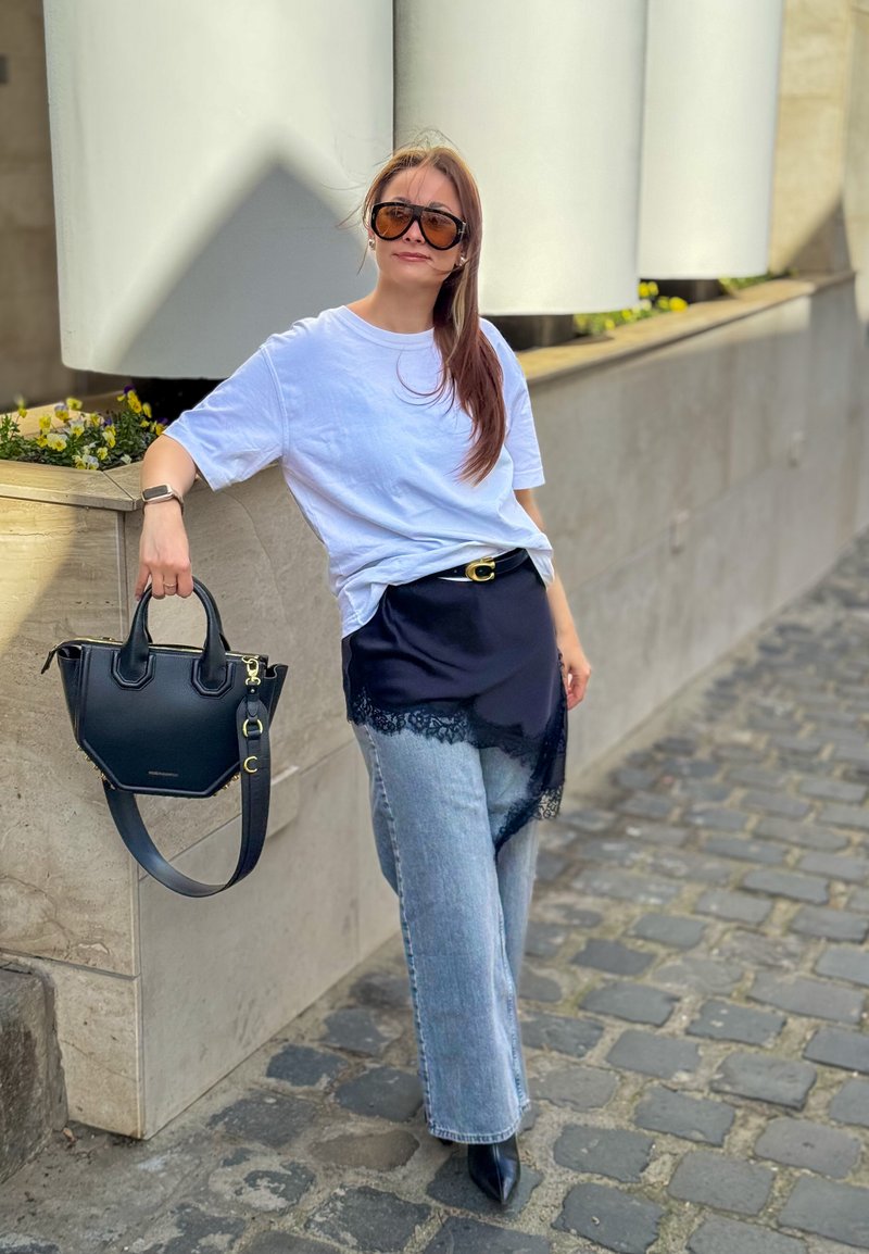 Femme portant un t-shirt blanc oversize, jupe en dentelle noire par-dessus un jean, chaussures noires pointues, lunettes de soleil et tenant un sac à main noir, appuyée contre un rebord en pierre.