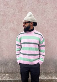 Ριγέ πουλόβερ σε ροζ, πράσινο και γκρι με στρογγυλή λαιμόκοψη, σε συνδυασμό με ένα ανοιχτό γκρι πλεκτό σκουφί. Σκούρο παντελόνι, στηρίζεται σε ένα υφασμένο ροζ τοίχο.