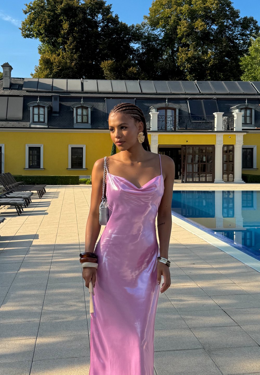 Abito di raso rosa con scollatura a cappuccio, caratterizzato da uno spacco vertiginoso. Accessoriato con una piccola borsa e bracciali, sullo sfondo di una piscina.