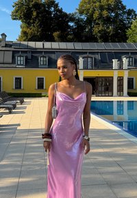 Abito di raso rosa con scollatura a cappuccio, caratterizzato da uno spacco vertiginoso. Accessoriato con una piccola borsa e bracciali, sullo sfondo di una piscina.