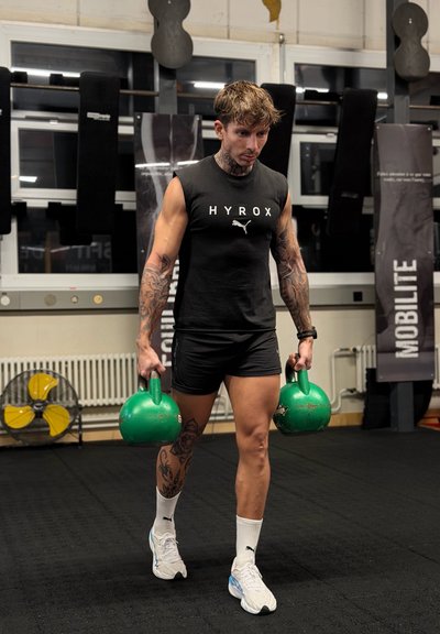 Hombre con tatuajes, vestido con ropa deportiva negra, lleva dos kettlebells verdes mientras camina en un gimnasio con colchonetas negras y pancartas de fitness.