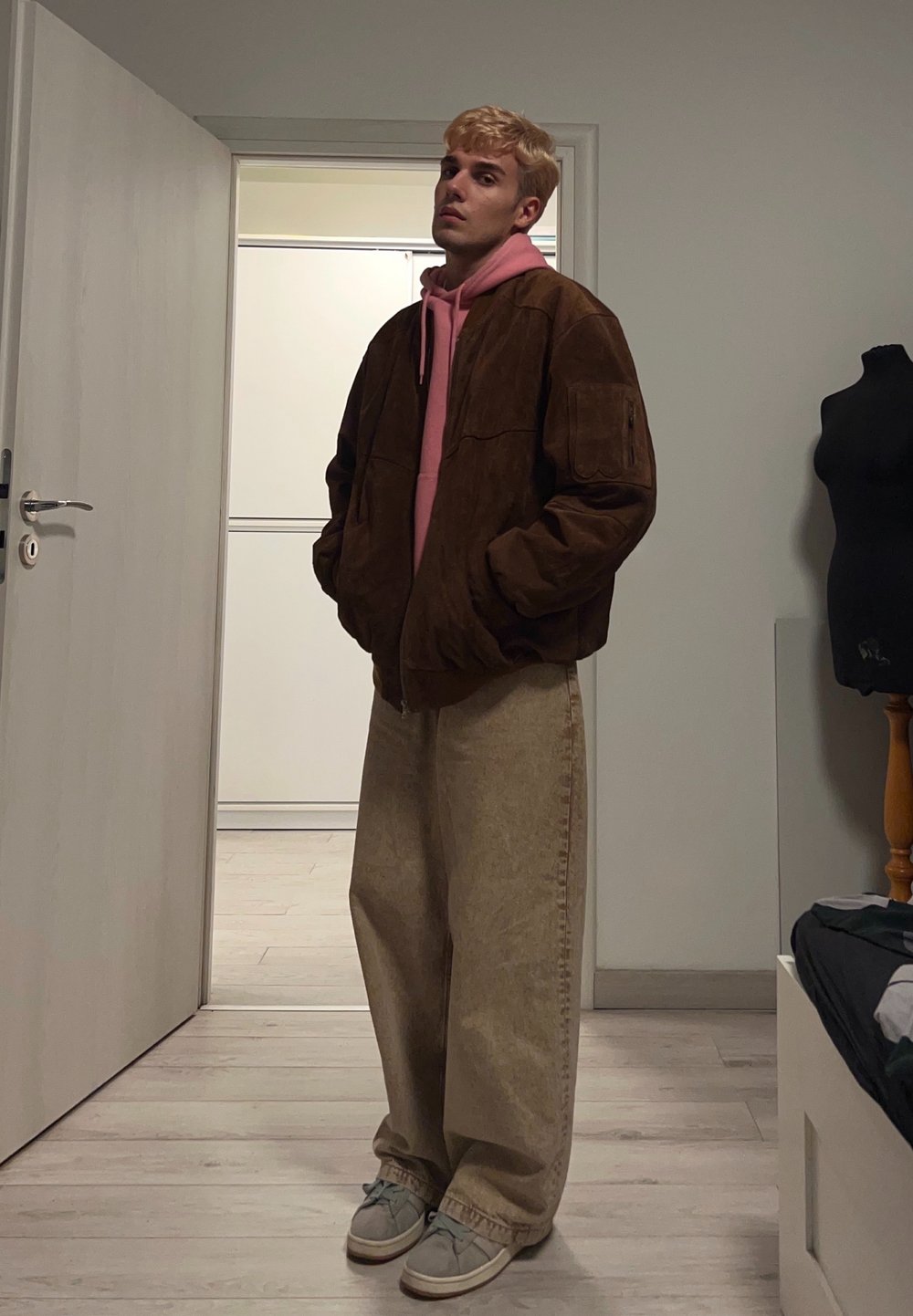 Veste oversize en daim marron superposée à un hoodie rose, associée à un pantalon large beige et des baskets grises. Fond intérieur minimaliste.