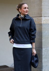 Femme aux cheveux blonds coiffés en chignon bas, portant un haut zippé gris foncé et une jupe, tenant une pochette noire tissée, avec un foulard à pois autour du cou.