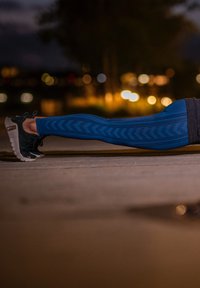 Kék sportos leggings, amelyet texturált cseppformájú minta díszít, fekete és szürke futócipővel párosítva, gyenge fényviszonyok között.