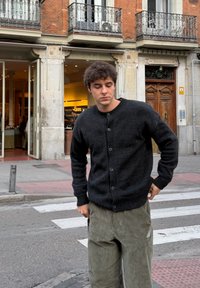 Zwarte gebreide cardigan met ronde hals en knoopsluiting, gecombineerd met groene corduroy broek. Grijzig textuur, smart-casual design.