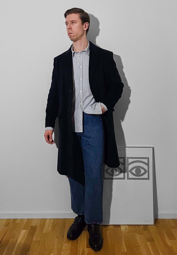 Long black coat over a white and blue striped shirt, paired with blue jeans and black shoes. Standing beside a framed graphic art piece.