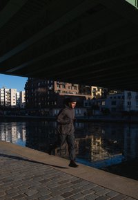 Homme athlétique portant une veste à capuche sombre et un pantalon, courant le long d'un chemin pavé près d'une surface d'eau réfléchissante sous un pont vert.