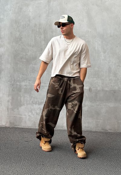 Camisa de manga corta de canalé beige, pantalones anchos de estampado de camuflaje en tonos oscuros, botas de trabajo en color caqui, gorra verde y blanca, gafas de sol, collares en capas.