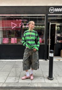 Grün-schwarz gestreiftes Langarmhemd, Camouflage-Cargo-Shorts, pinke Schuhe, weiße Socken, goldene Kettenhalskette. Urbaner Hintergrund.