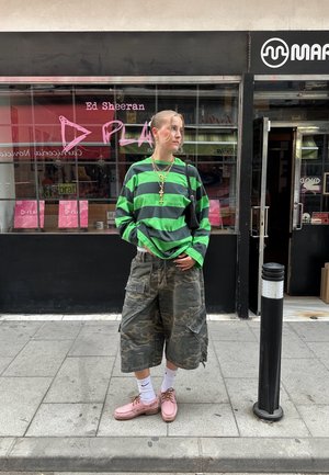 Grön och svart randig långärmad skjorta, kamouflage cargo-shorts, rosa skor, vita strumpor, guld kedja halsband. Urban miljö i bakgrunden.