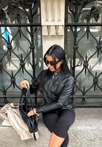Giacca di pelle nera con dettagli zip, borsa nera e trench beige su una superficie di pietra, accompagnati da uno sfondo decorativo in ferro battuto.