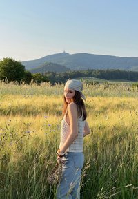 Háčkovaný top na ramínka v of-white barvě, vysoké modré džíny, držící malou hnědou tkanou tašku, stojící na travnatém poli s divokými květinami.