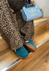 Tirkizni semiš športni čevlji z rjavo gumo, v kombinaciji z širokimi hlačami z leopardjim vzorcem in svetloblujim ročajem, ki ima zlati logo.
