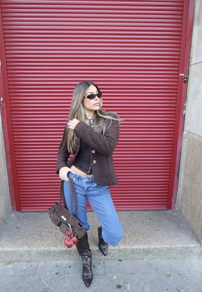 Mujer con gafas de sol, chaqueta marrón, top negro, vaqueros de pernera ancha y botas puntiagudas, sosteniendo un bolso marrón frente a un fondo de persiana roja.