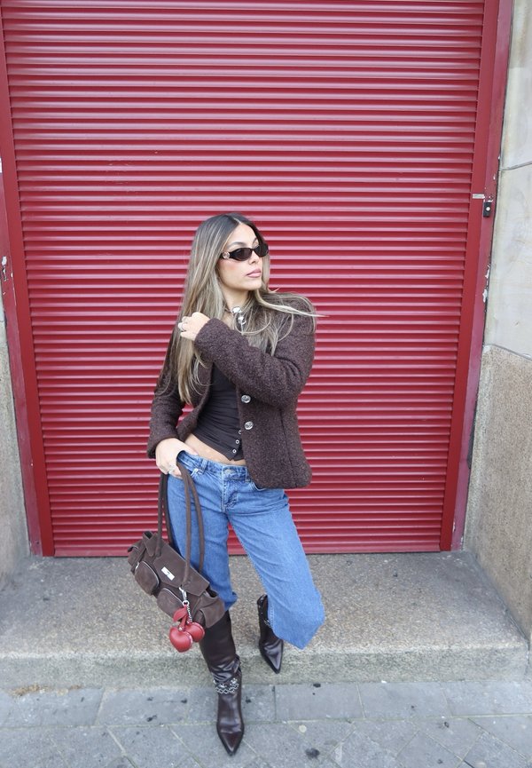 Nainen, joka käyttää aurinkolaseja, ruskeaa takkia, mustaa toppia, leveälahkeisia farkkuja ja teräväkärkisiä saappaita, pitää ruskeaa käsilaukkua punaisen sammutetun ikkunaluukun edessä.