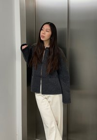 Veste en laine gris foncé avec de grandes manches, fermeture à crochets sur le devant et deux poches, associée à un pantalon de couleur crème sur un fond métallique.