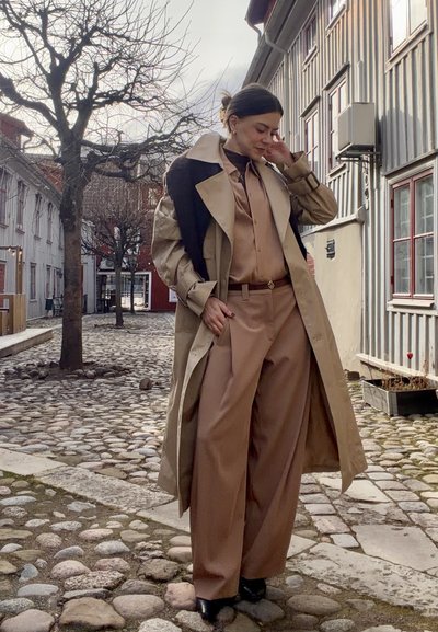 Mujer con pantalones anchos color marrón claro, blusa a juego y gabardina, con un suéter negro sobre los hombros, de pie en una calle empedrada.