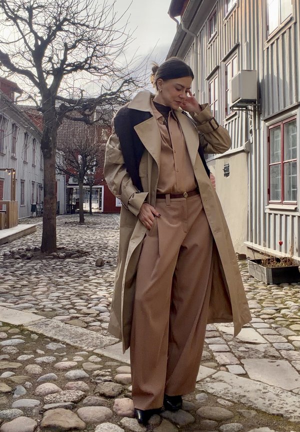 Žena v hnědě zbarvených širokých kalhotách, odpovídající blůze a trenčkotu s černým svetrem přehozeným přes ramena stojí na dlážděné ulici.