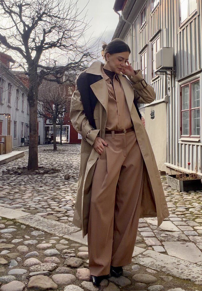 Kvinde i tan-farvede vidde bukser, matchende bluse og trenchcoat med sort sweater draperet over skuldrene, stående på en brostensbelagt gade.