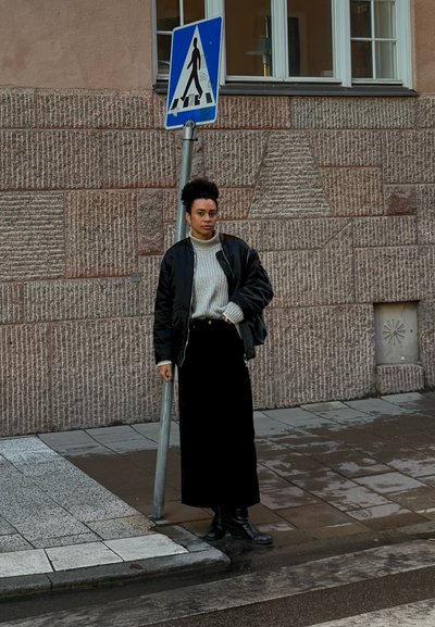Chaqueta bomber negra sobre un suéter de cuello alto gris, combinado con una falda larga negra y botas negras. De pie junto a una señal de peatones al lado de paredes de piedra texturizada.